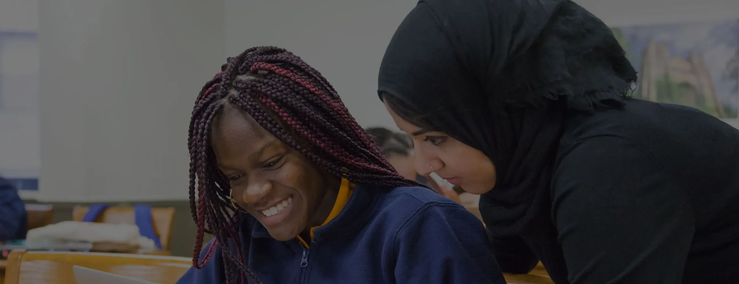 A Success Academy teacher leans over a high school female student who is smiling while looking at a laptop screen.