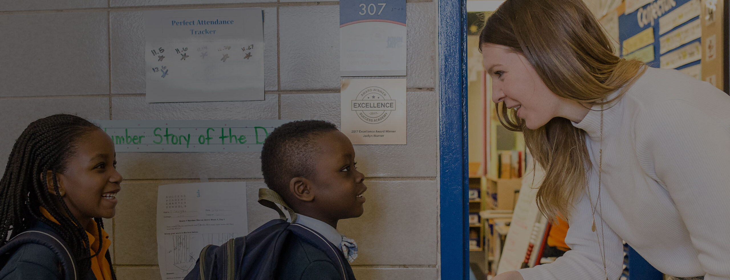 A smiling elementary school teacher in a light-colored sweater greets two young SA elementary school students carrying backpacks.