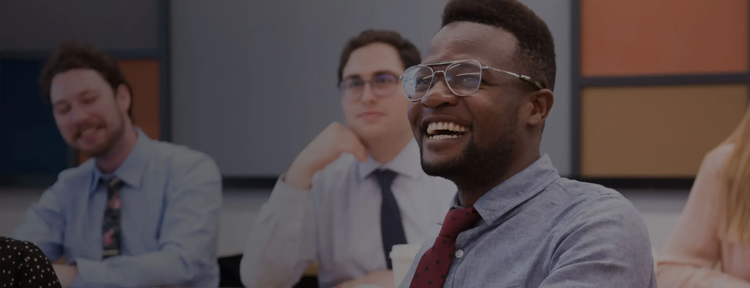 A SA employee in a blue shirt and red tie, wearing glasses, smiles broadly while sitting in a meeting. Other employees are visible in the blurred background.