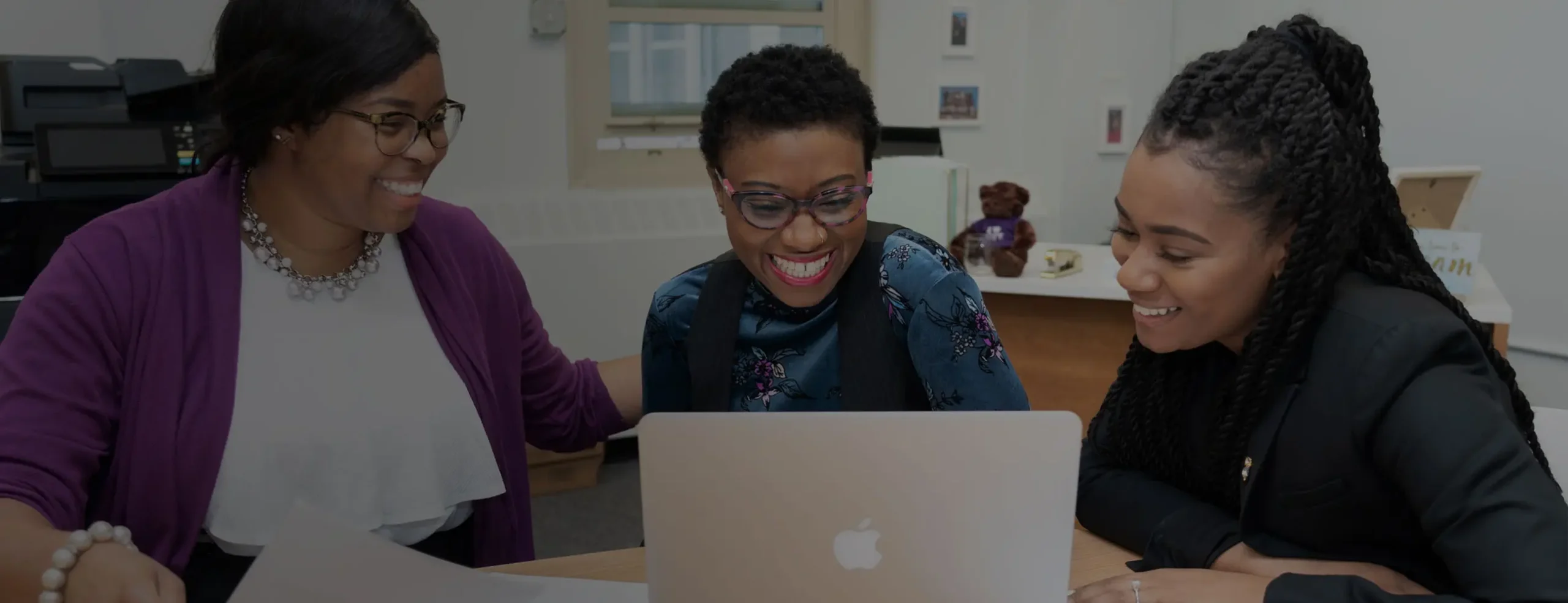Three women are smiling and looking at a laptop screen together around a wooden table in an office. One woman wears glasses and a purple cardigan, another has short curly hair and glasses, and the third has long braided hair.