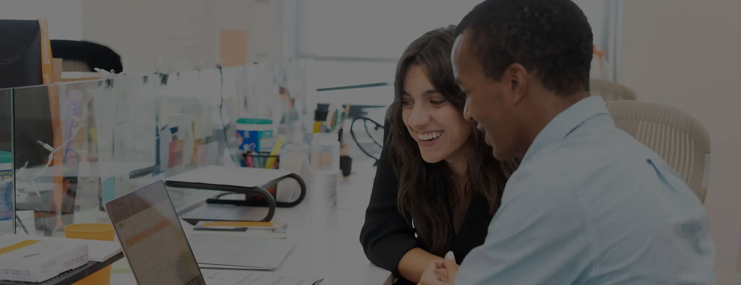 A woman with dark hair and a man in a light blue collared shirt are smiling and looking at a laptop screen together in the SA network office.