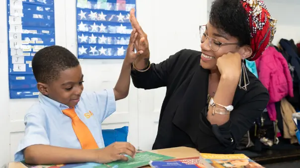 A elementary school male student and a female teacher, are high-fiving across a table in a classroom, both smiling. The teacher wears glasses and a head wrap.