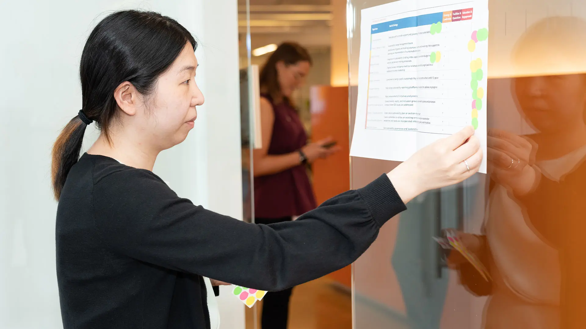 A female SA network employee with dark hair pulled back in a ponytail is placing a document with blue and pink sections on a clear partition in the SA network office. Another employee is visible in the background.