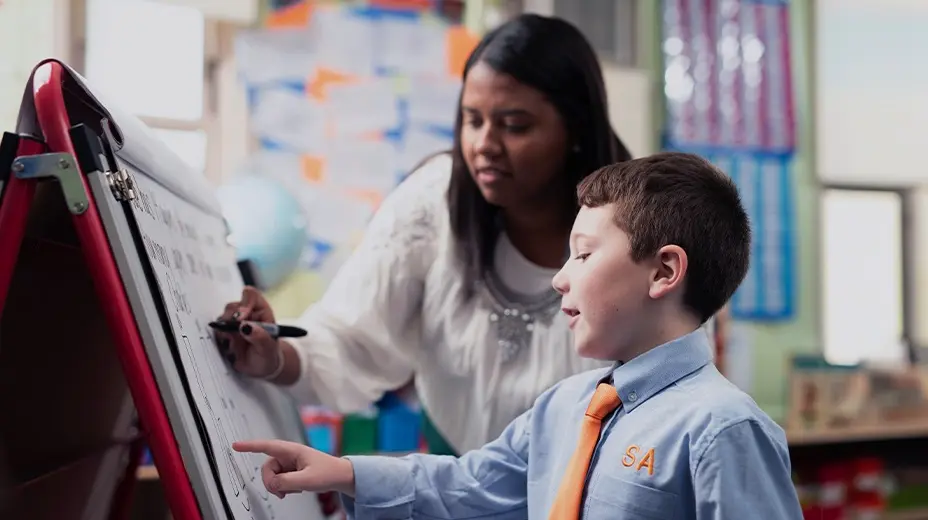 An elementary school male student in a blue shirt with an orange tie points at a whiteboard while a female teacher, writes on it behind him in a classroom setting.