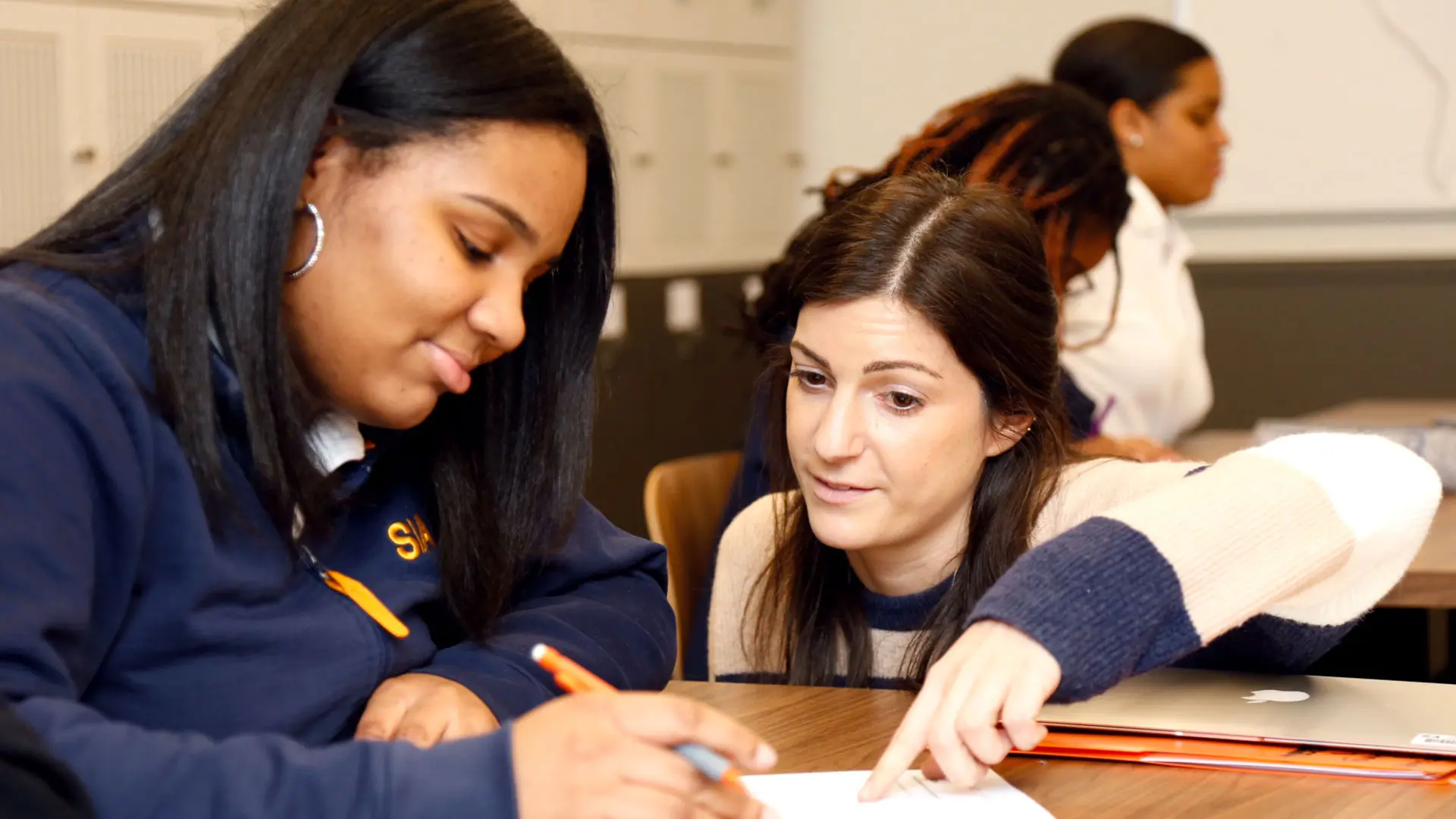A female teacher with long brown hair, wearing a striped sweater, points to a document while helping a female high school student with long dark hair, wearing a navy hoodie, in a classroom.