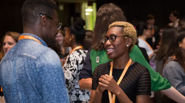 A woman with short, blonde hair and glasses smiles while talking to a man wearing a denim shirt and glasses. Both are wearing orange lanyards, and other people are in the blurred background.