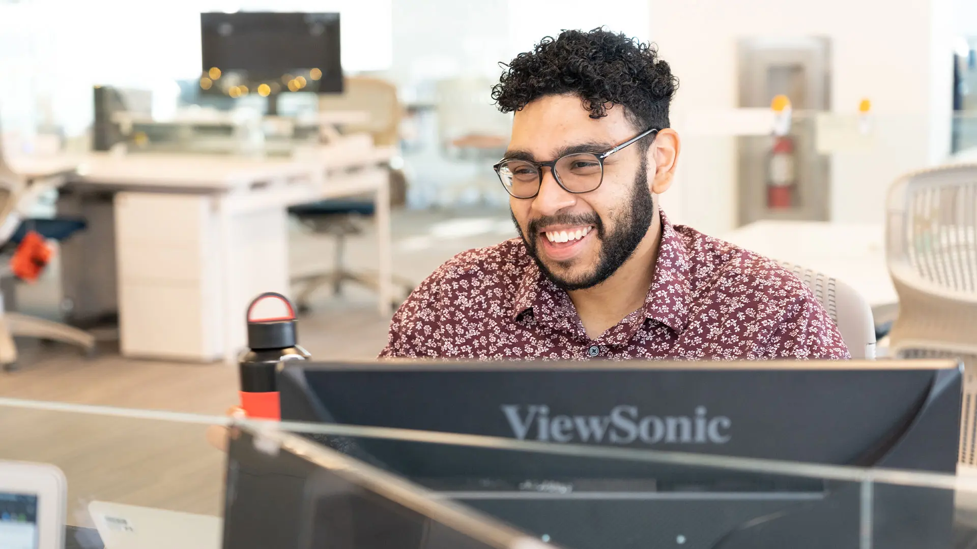 A smiling man with curly dark hair and glasses is looking at a computer monitor in the SA network office.