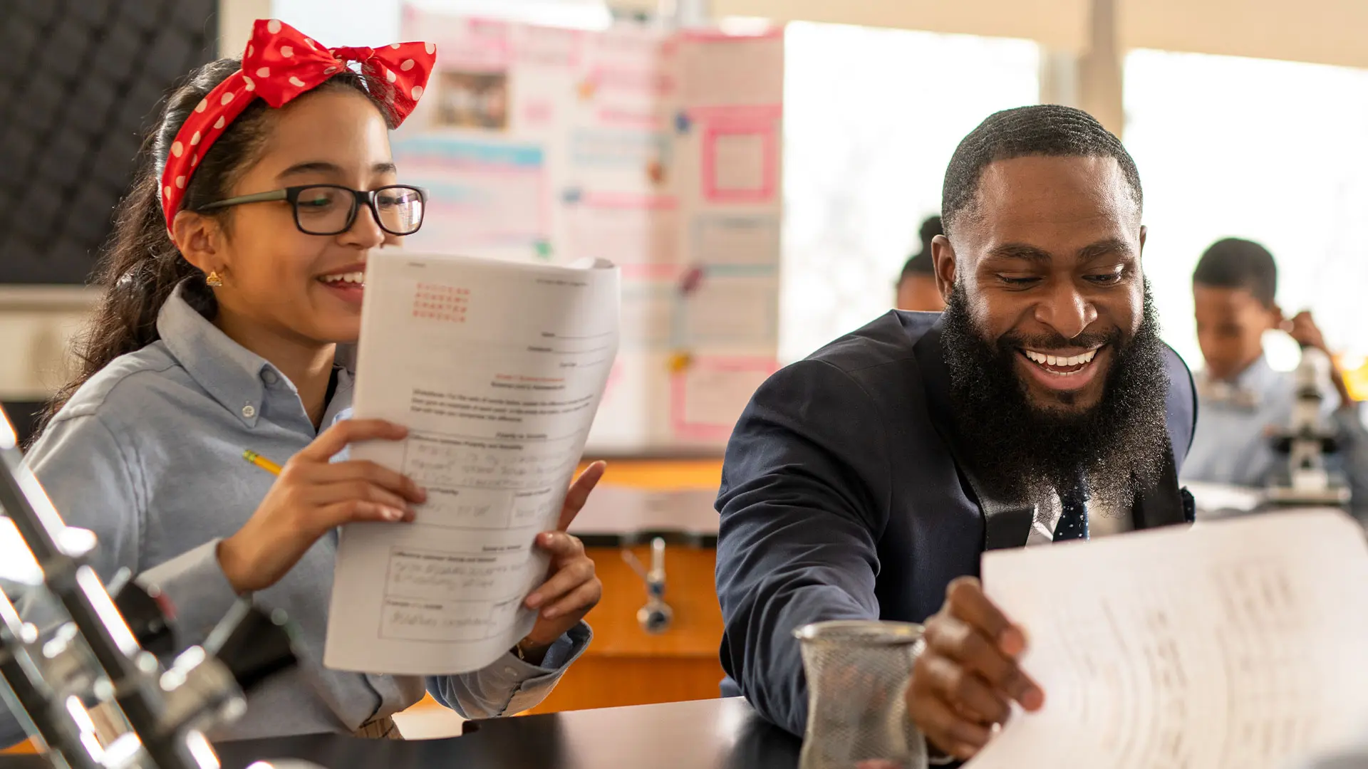 A smiling middle school girl with glasses and a red polka-dot headband holds a paper while a teacher with a beard leans in, looking at a paper in a classroom.