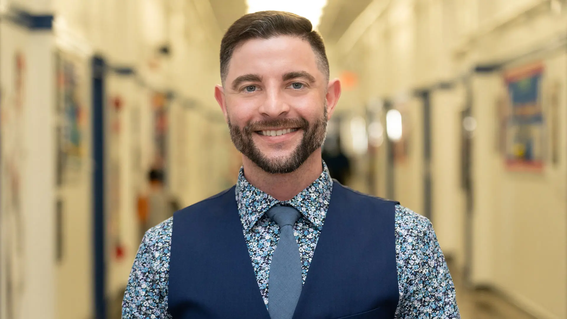 An SA male employee with short hair and an orange tie smiles at the camera in a school hallway with lockers.