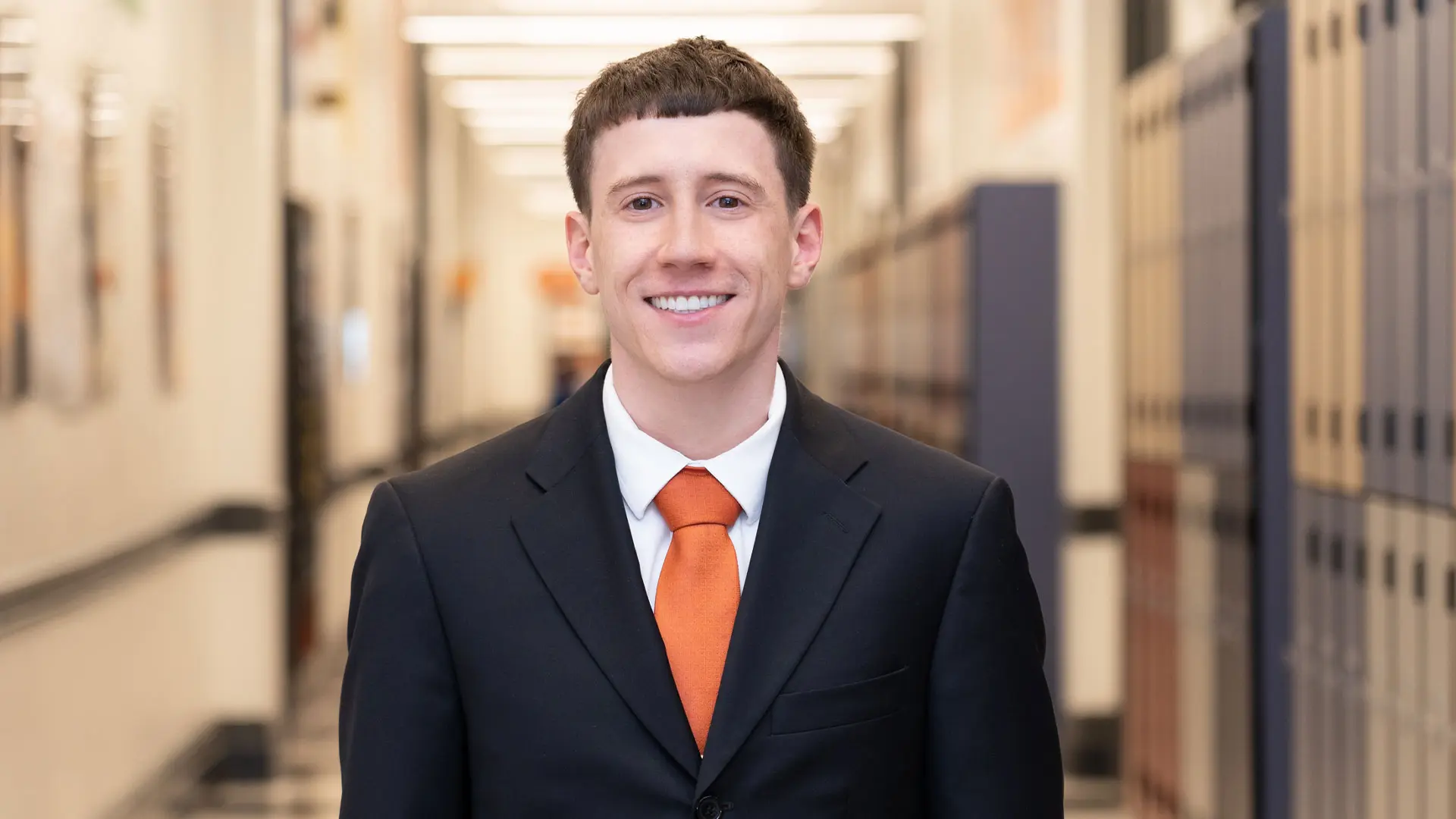An SA male employee with a dark suit and orange tie smiles directly at the camera in a school hallway with blue lockers.
