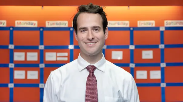 A smiling male teacher wearing a white collared shirt and a red tie stands in front of an orange and blue bulletin board with days of the week labeled.