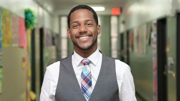 A smiling male SA teacher wearing a white shirt, a grey vest, and a colorful plaid tie stands in the middle of a school hallway with lockers and artwork on the walls.