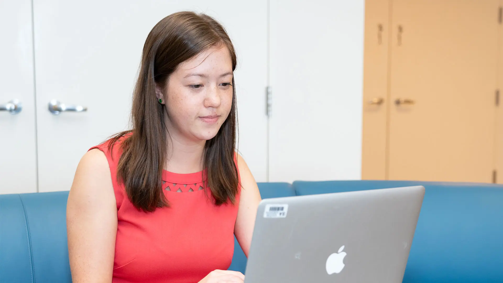 A young SA employee with brown hair, wearing a red top, sits on a blue couch or bench and looks at a laptop. In the background, there is a white wall with doors or cabinets.