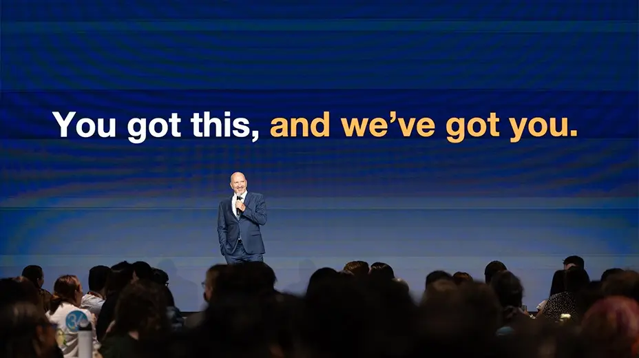 A man in a suit stands on a stage addressing a crowd, with the words "You got this, and we've got you." displayed in orange and white text on a blue screen behind him.