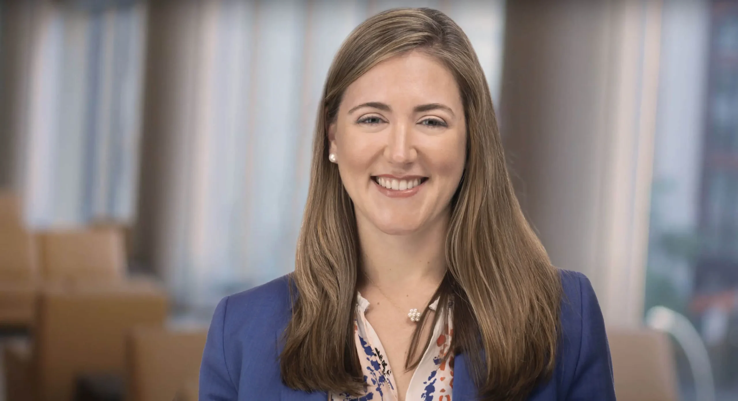 A smiling woman with long brown hair, wearing a blue blazer over a patterned blouse, looks directly at the camera. The background appears to be an office or professional setting with blurred furniture.