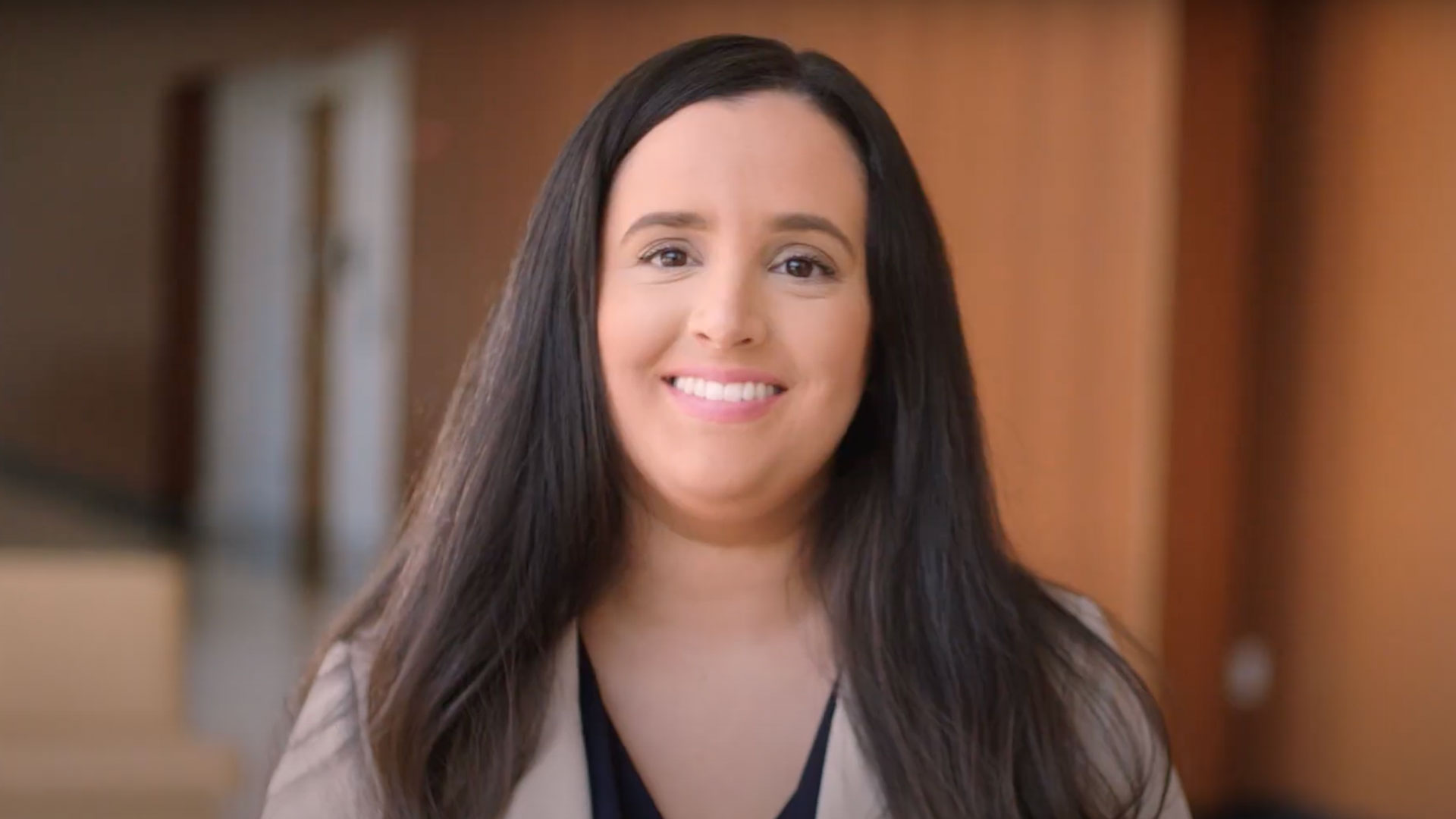A smiling SA teacher with long, dark hair looks directly at the camera, wearing a light-colored blazer over a darker top. The background is a blurred school setting.