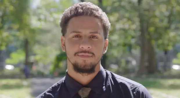 A young male teacher with light brown curly hair and a beard, wearing a dark collared shirt and tie, looks directly at the camera with a neutral expression. The background is a blurred outdoor setting with green trees.