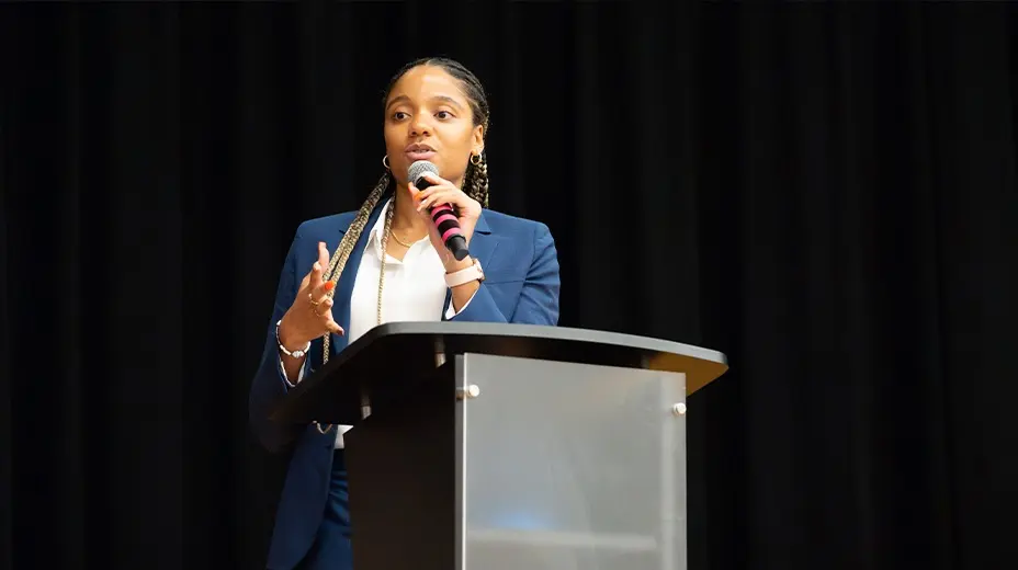 A young woman with braided hair, wearing a dark suit and a white blouse, stands at a podium holding a microphone and speaking, with a dark background behind her.