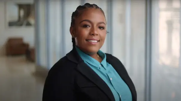 A young female SA employee with braided hair and a bright smile looks directly at the camera, wearing a black blazer over a teal collared shirt. The background is the SA network office.