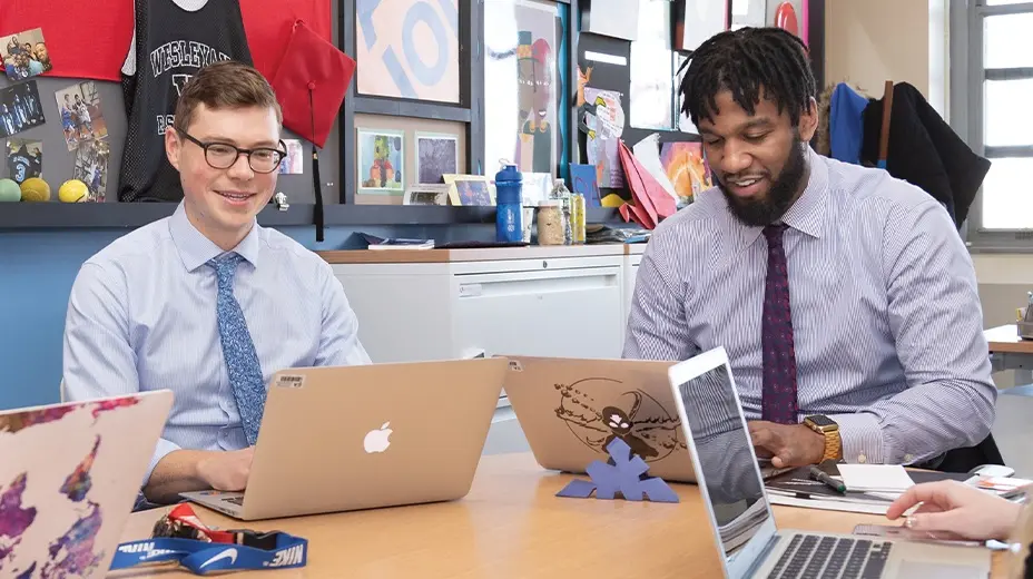 Two SA employees are seated at a table, each working on a laptop. The man on the left, wearing glasses and a light blue shirt with a patterned tie, looks up and smiles. The man on the right, with dark skin and a beard, wearing a striped shirt and dark tie, looks down at his laptop with a slight smile. The background is a classroom setting.