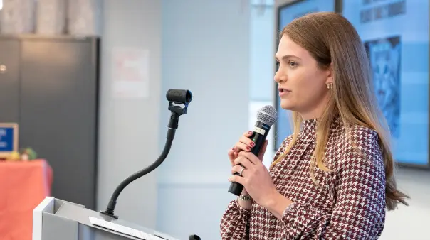 A young woman with long, light brown hair, wearing a patterned high-neck top, stands at a podium holding a microphone and speaking. The background is a brightly lit indoor space with a light-colored wall and what appears to be framed pictures or screens.