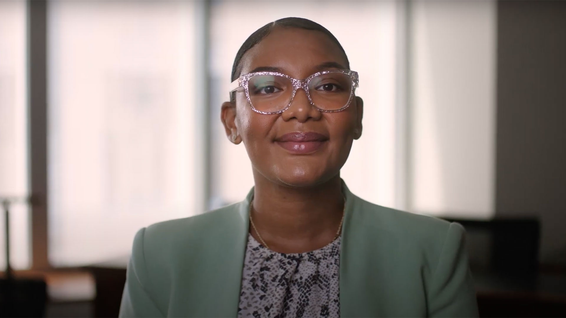 A smiling female SA employee, wearing large, clear-framed glasses and a light green blazer over a snake-print top, looks directly at the camera. The background is the SA network office.
