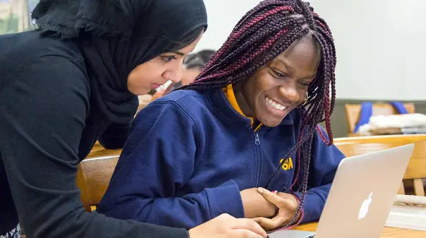 A teacher wearing a black head covering is helping a high school student with braided hair, who is smiling and looking at a laptop screen.