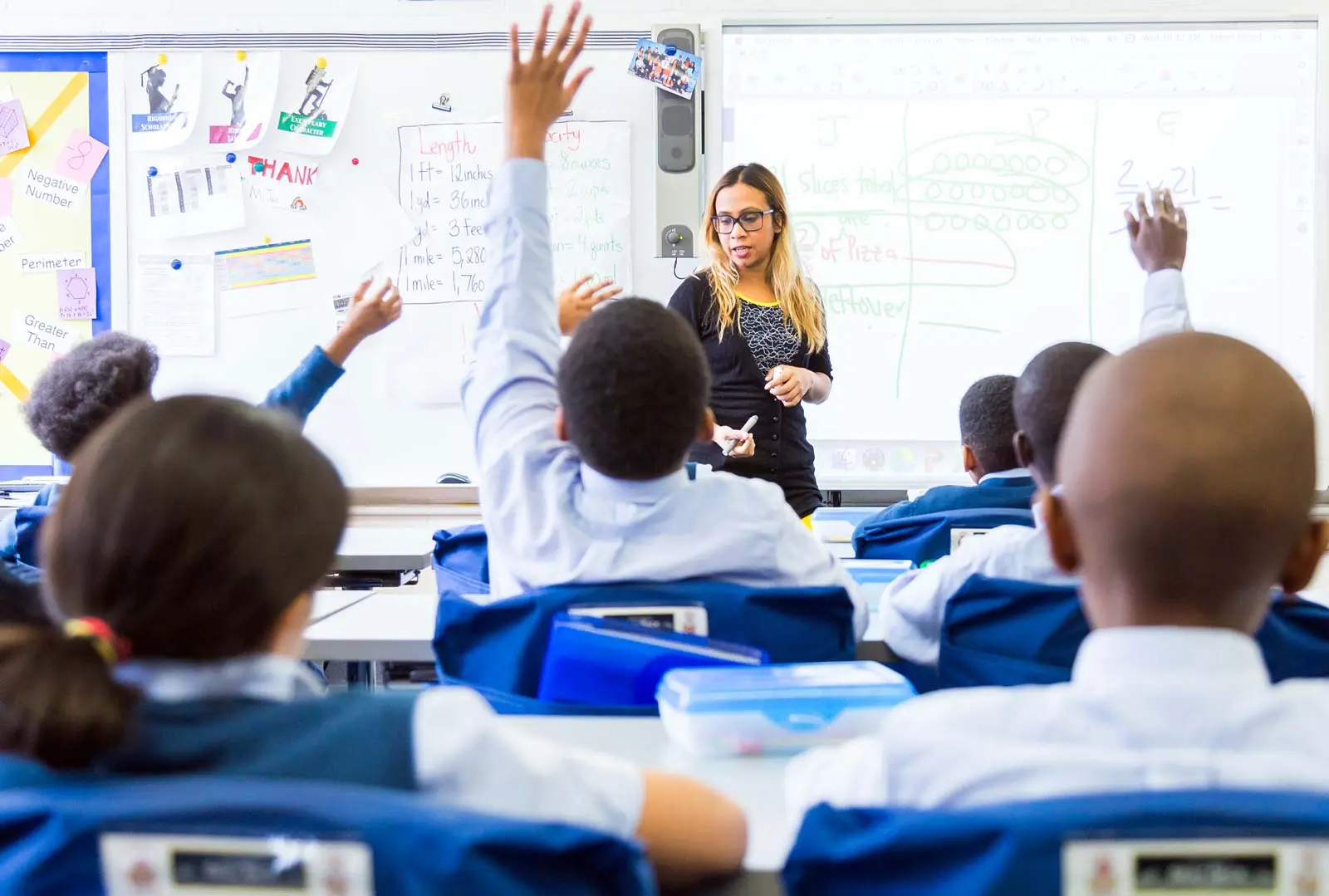 A bright classroom with a teacher standing in front of a whiteboard, facing a elementary school students seated at desks. Several students have their hands raised.