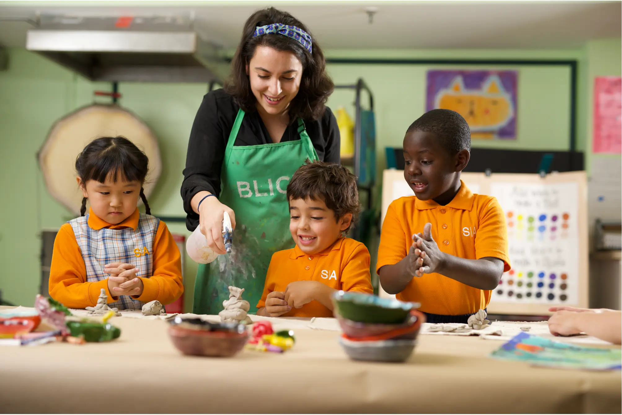 A elementary school female teacher smiles as she helps an elementary school students with an art project at a wooden table in a classroom. The students are wearing SA uniforms.