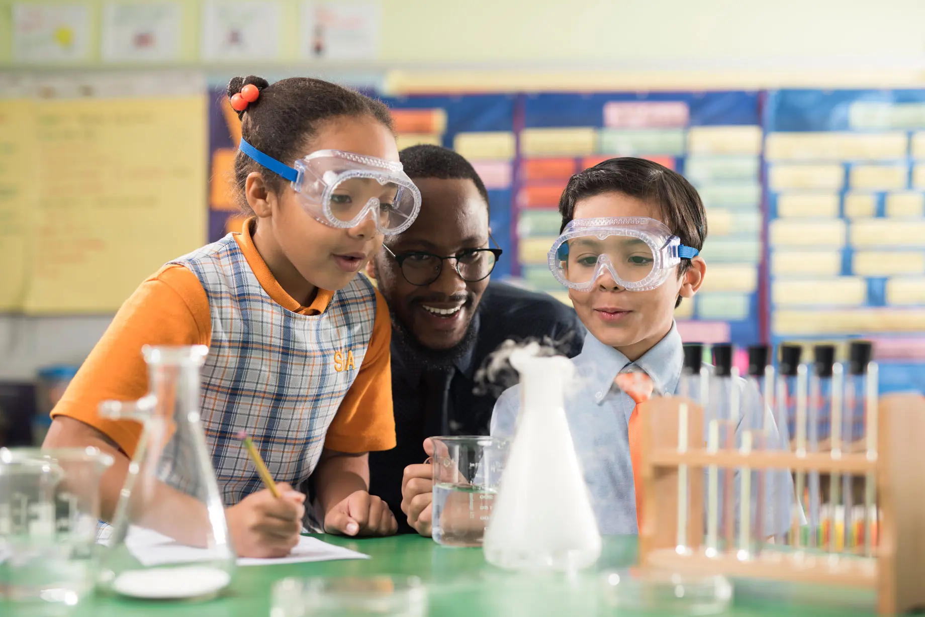 Two elementary school students are wearing goggles andare engaged in a hands-on science activity, looking at materials on a table with a tacher observing.