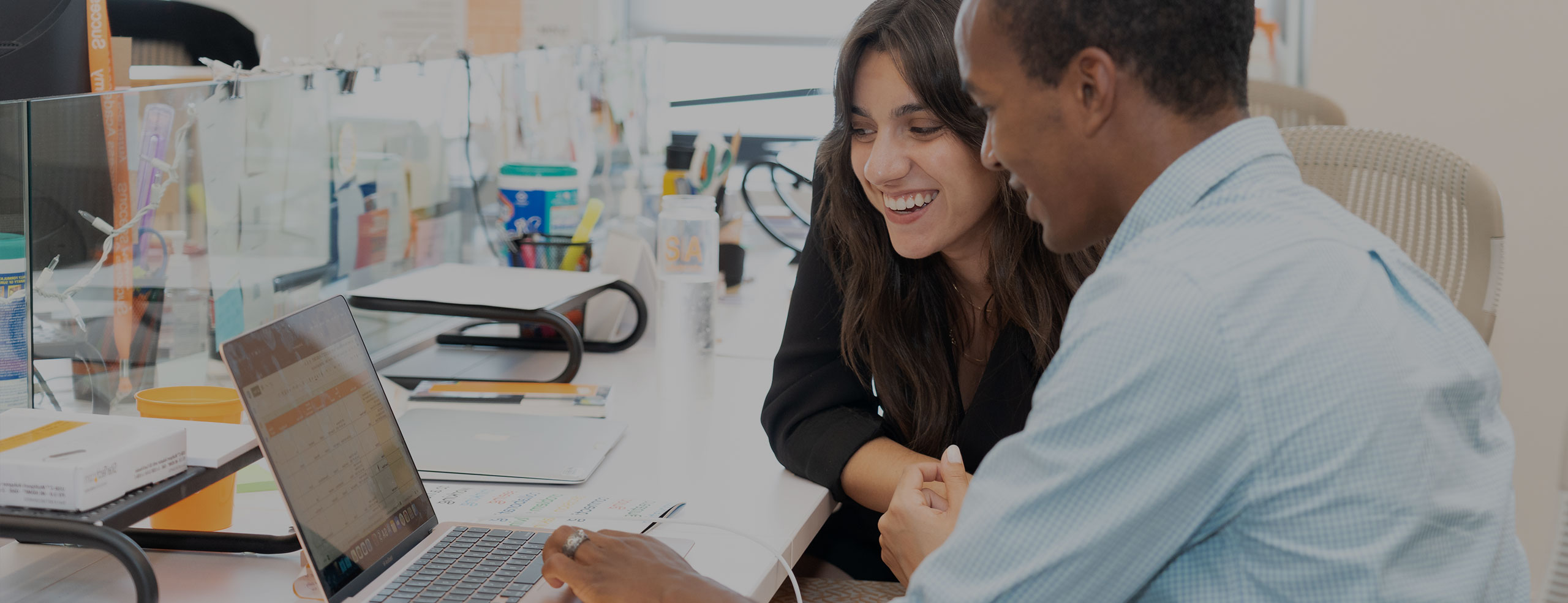 Two SA network colleagues, a man and a woman, are smiling and collaborating at a desk in the SA network office, looking at a laptop screen.
