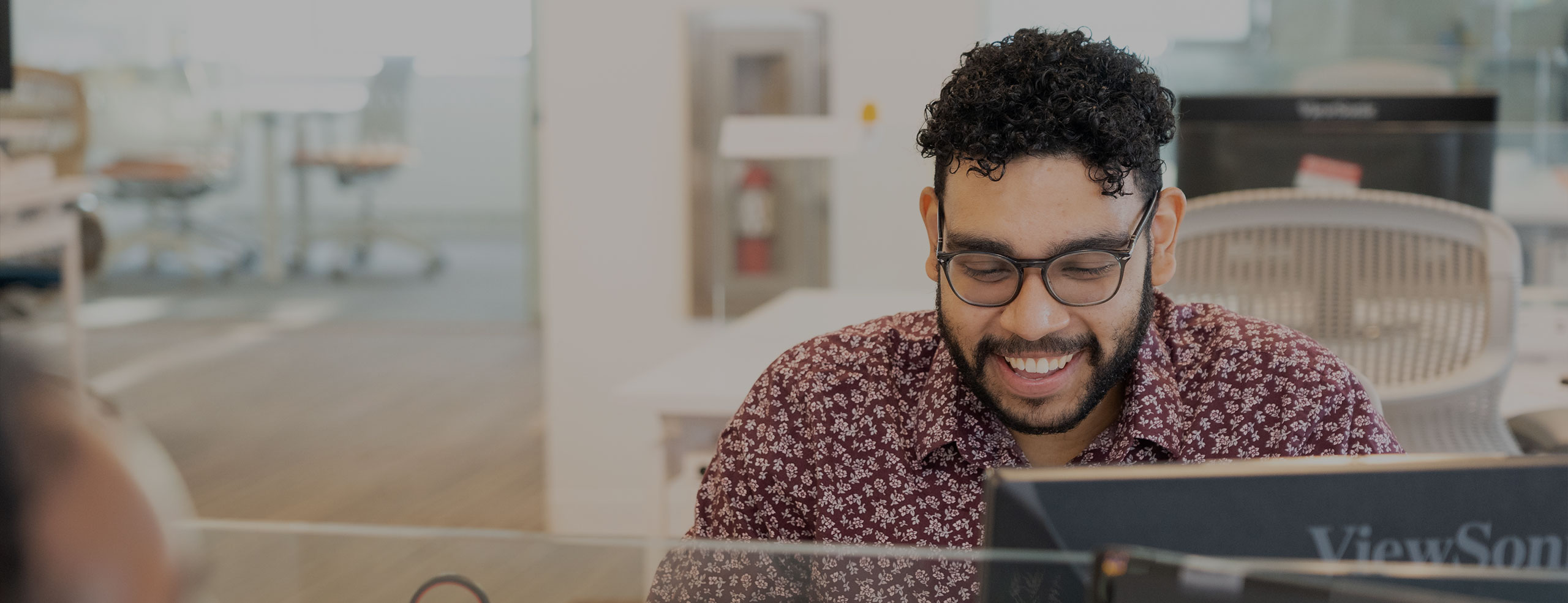 A smiling man with glasses and curly hair is looking down at a laptop in the SA network office.
