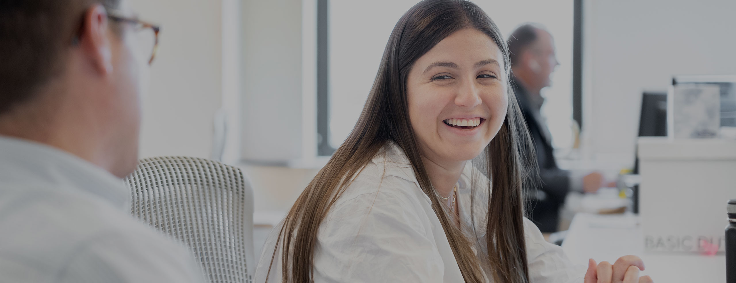 A smiling female SA employee with long brown hair looks over her shoulder while sitting at a desk in the SA network office.