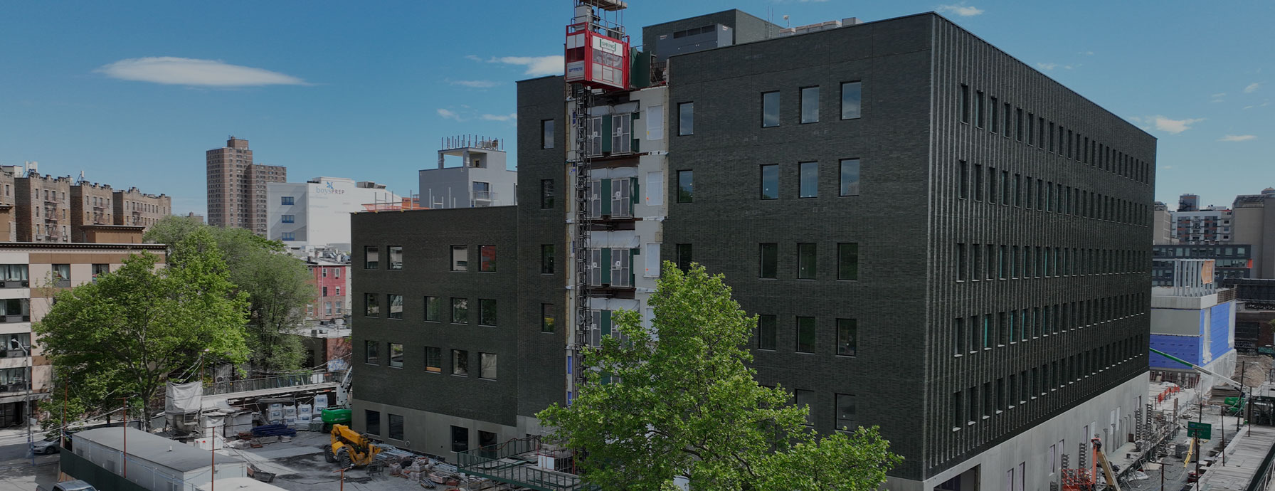An aerial rendering of Success Academy's Bronx K-12 campus. The campus will be opening in Fall 2026. The design shows a large, dark-colored building under construction in an urban environment, with some trees and other buildings visible. A construction elevator is on the side of the building.