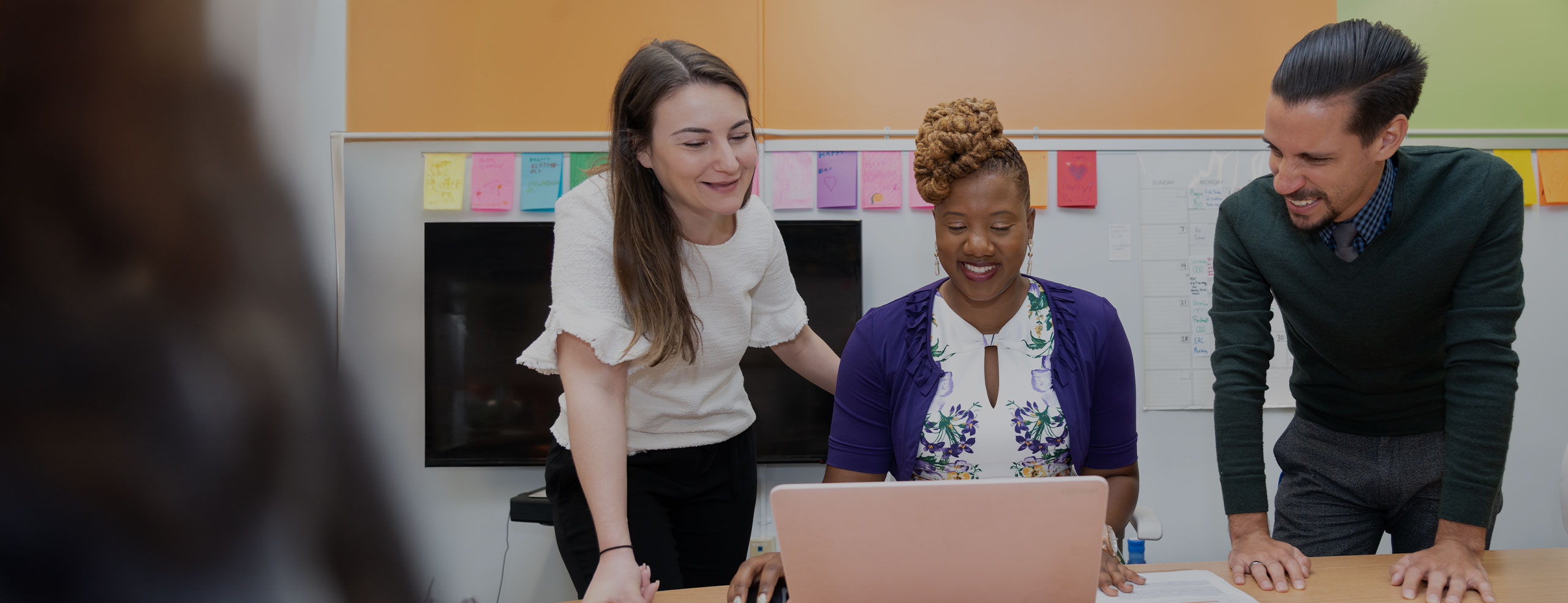 Three SA network colleagues are gathered around a laptop in the SA office. A woman in a white shirt stands next to a woman in a purple top who is looking at the laptop. A man stands on the right, also looking at the laptop and smiling.