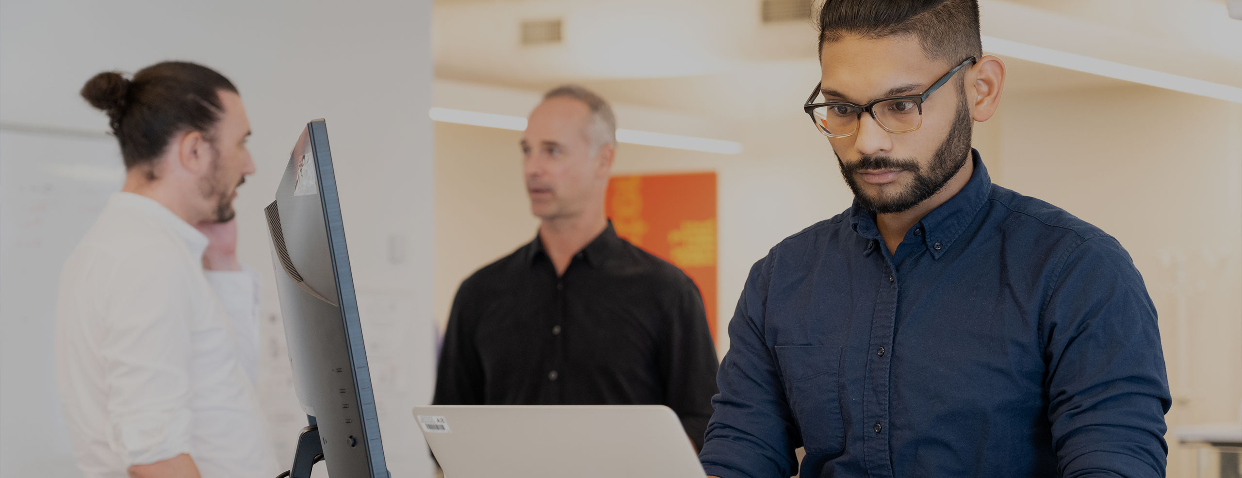 An SA network employee with a beard and glasses is intently looking at a computer monitor in a SA network conference room. Two other men are visible in the blurred background.
