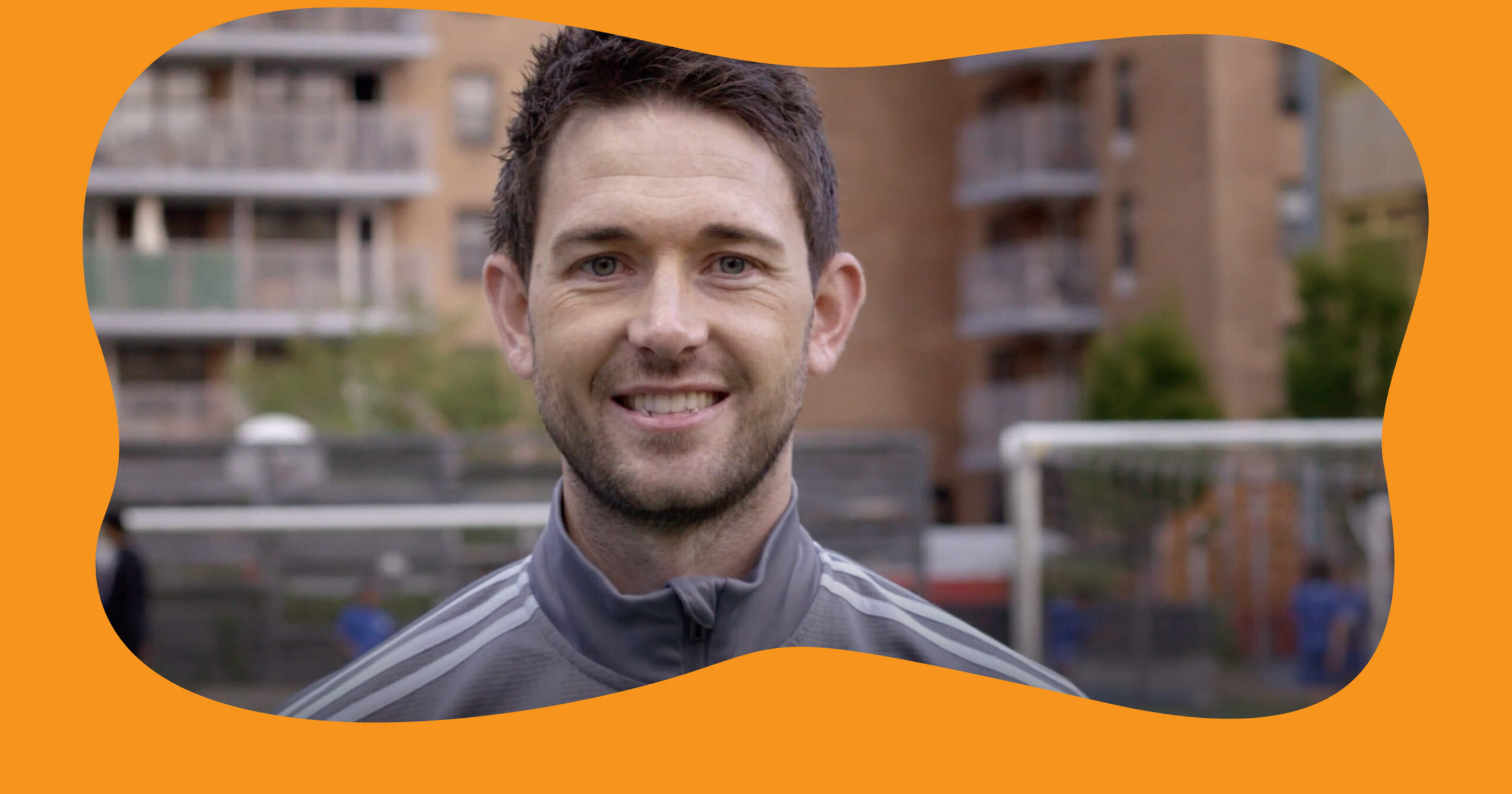 A smiling soccer coach in a grey athletic jacket with white stripes on the shoulders smiles at the camera. He is outdoors on a soccer field, with blurred apartment buildings and soccer goals in the background.