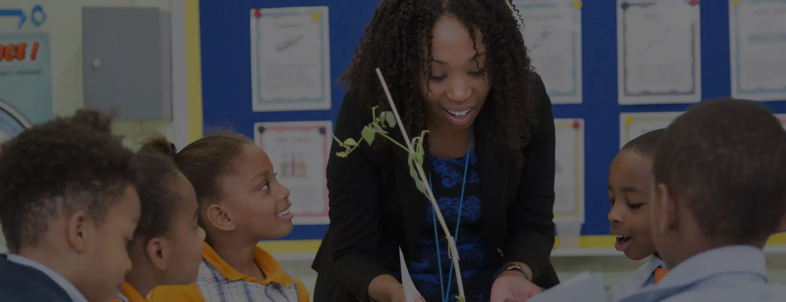 A Black female SA teacher with long, dark curly hair, wearing a blazer, smiles and leans over a table to interact with a group of diverse elementary school students. She is holding a small plant, and the students are looking at it with interest.