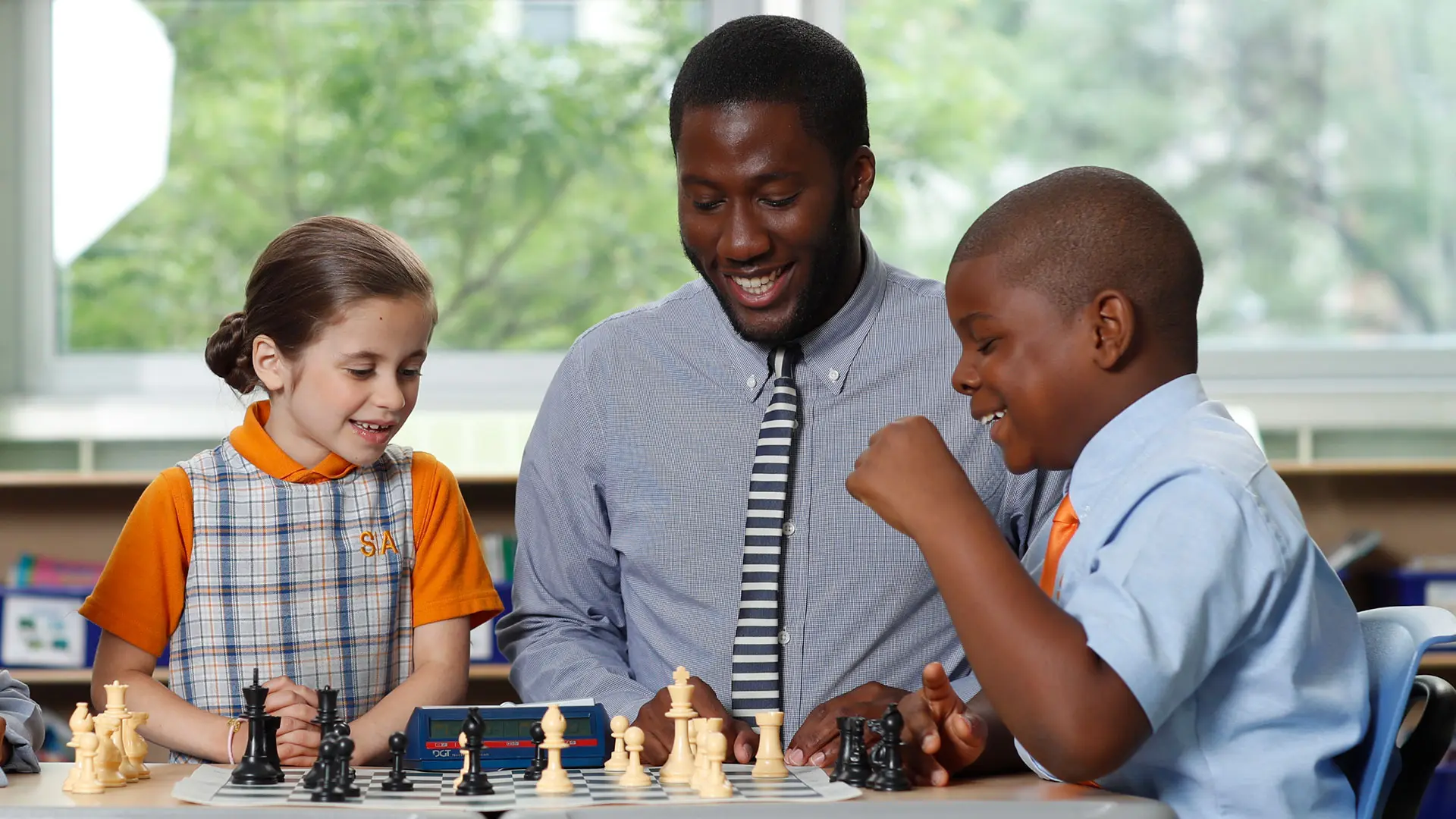 A two elementary school students are engaged in a chess game while a chess teacher observes and smiles.