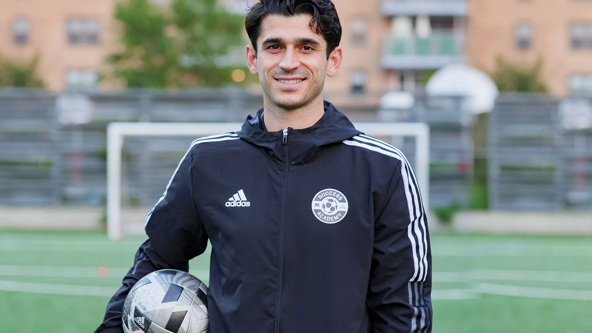 A smiling male coach with dark hair, wearing a black Adidas jacket with "Success Academy" on a circular logo, holds a soccer ball under his arm. He stands on a green turf field with a white soccer goal in the background.